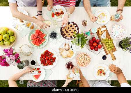 Giovani studenti aventi un pasto al di fuori, bere e mangiare la torta, anguria, mele e uva ad un tavolo con fiori Foto Stock