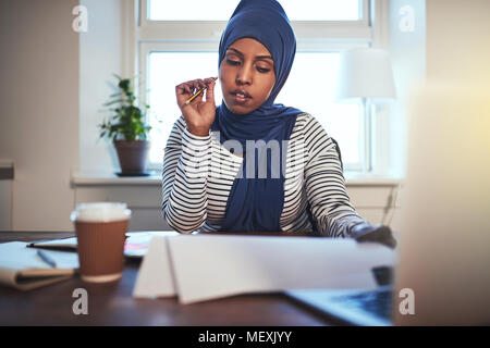 Focalizzato giovani arabi imprenditore femmina indossando un hijab seduti ad una scrivania nel suo ufficio a casa attraverso la lettura di documenti Foto Stock