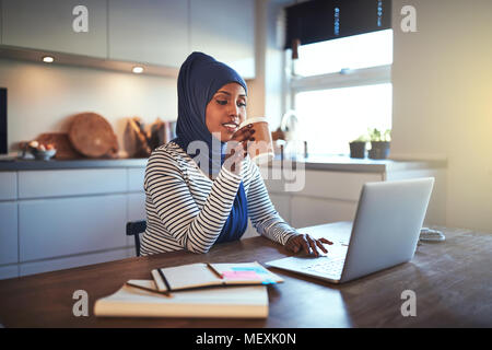 Giovani arabi imprenditore femmina indossando un hijab a bere un caffè e lavorare su un laptop mentre si siede a un tavolo nella sua cucina Foto Stock