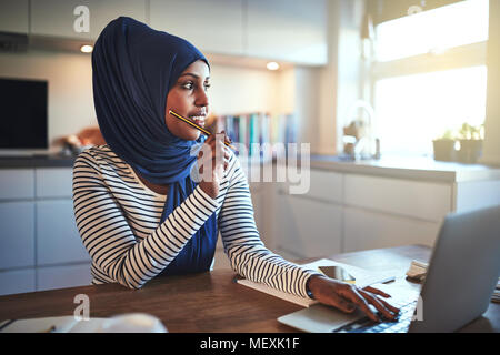 Giovani arabi imprenditore femmina indossando un hijab a guardare in profondità nel pensiero mentre è seduto al suo tavolo da cucina al lavoro su un notebook Foto Stock