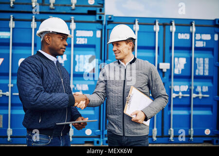 Due ingegneri sorridente indossando hardhats in piedi da contenitori di spedizione su un dock commerciale agitando mani insieme Foto Stock