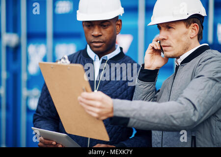 Freight manager parlando al cellulare e la lettura di un elenco di inventario su un clipboard mentre in piedi con un collega in una spedizione commerciale cantiere Foto Stock