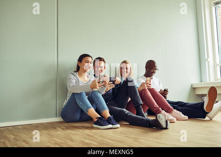 Gruppo di diversi giovani colleghi seduti insieme su un pavimento di Office utilizzando i loro telefoni cellulari durante una pausa in un ufficio moderno Foto Stock