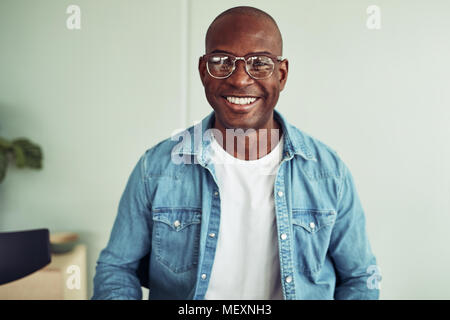 Casualmente vestito giovane imprenditore africano con gli occhiali e sorridente con fiducia mentre è seduto da solo in un ufficio Foto Stock