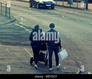 La popolazione locale coppia giovane uomo e donna shopping spingendo la PRAM su strada marciapiede marciapiede del traffico su strada attraversando Castlemilk, Glasgow, Regno Unito Foto Stock