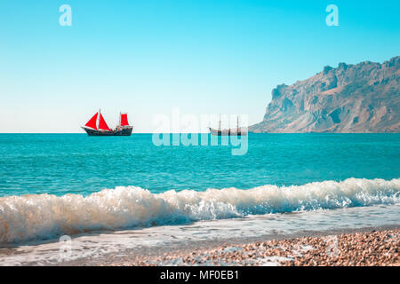 Due navi andare dal mare all'orizzonte. Uno di loro con vele di rosso. Onda in primo piano. La montagna è sulla destra. Cielo limpido e mare turchese. Foto Stock