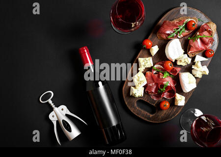 Delizioso antipasto al vino - prosciutto, formaggio, fette di baguette, pomodori, servita su una tavola di legno e vetro con un bicchiere di vino rosso sulla superficie nera Foto Stock