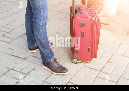 Uomo in abiti casual in piedi con la valigia rossa in corrispondenza della stazione. Giovane viaggiatore moderno in attesa di viaggio dealyed. Persona con bagaglio Viaggio di partenza. Foto Stock