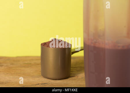 La proteina del siero di latte integratore alimentare per la formazione e l'esercizio. Close up di scoop con il cioccolato in polvere e shaker su tavola di legno. Colore di sfondo: giallo. Foto Stock