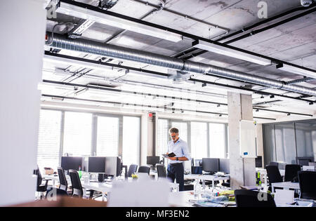 Imprenditore maturo in piedi in un piano aperto ufficio notebook di contenimento Foto Stock