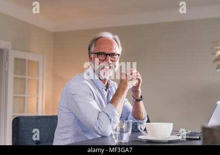 Ritratto di felice uomo maturo con il computer portatile e la tazza di caffè seduti al tavolo della sua stanza vivente Foto Stock