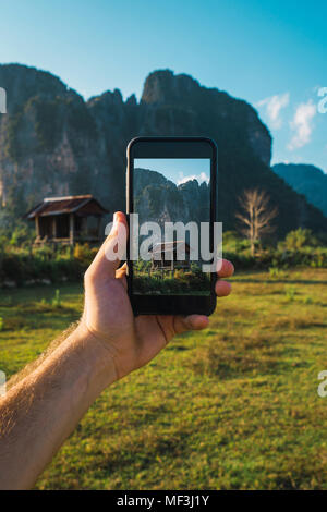 Laos, mano tenendo lo smartphone, Display con capanna in legno e montagne Foto Stock