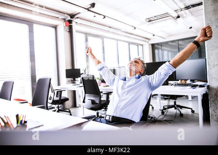 Happy businessman maturo seduto alla scrivania in ufficio solleva le braccia Foto Stock