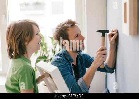 Figlio guardando padre guida un chiodo nel muro a casa Foto Stock