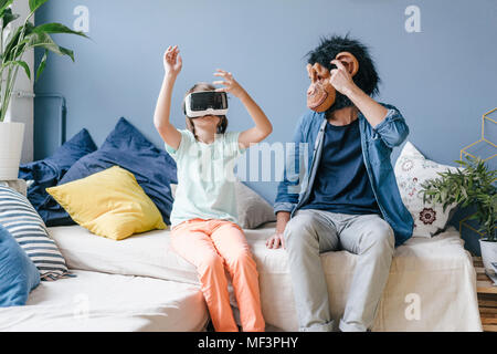 Padre indossa la maschera di scimmia guardando il figlio che indossa gli occhiali VR a casa Foto Stock