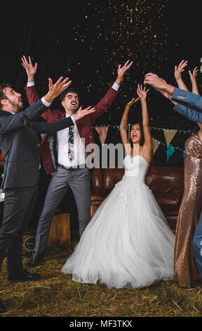 Allegro sposi e amici alzando le braccia mentre i confetti cade sopra le loro teste in una notte party all'aperto Foto Stock