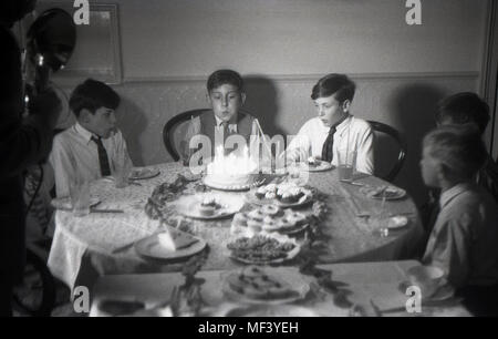 1948, storico, giovane ragazzo soffiando fuori le candeline sulla sua torta di compleanno come egli ha un compleanno tea party con i suoi amici Foto Stock