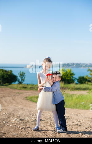 Junior fratello abbracciare sorella in abito bianco azienda papavero rosso bouquet sulla costa del mare sullo sfondo Foto Stock