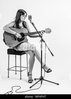 Foto di una giovane donna con i capelli lunghi seduto su uno sgabello suonando una chitarra acustica su sfondo bianco. Foto Stock