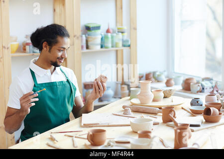 Giovani razza mista potter maschile seduto al tavolo del workshop, l'applicazione di smalto sulla artigianale di pentola di creta con pennello e sorridente Foto Stock