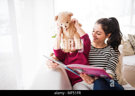 Giovane madre e una piccola ragazza con Teddy bear a casa, la lettura di un libro. Foto Stock