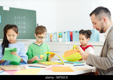 I bambini della scuola elementare seduta in Aula e rendendo origami con insegnante maschio pur avendo dopo la scuola tecnica di classe Foto Stock