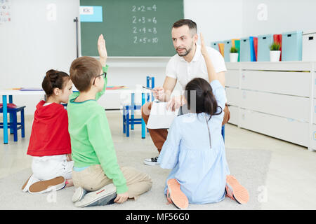Studenti della scuola primaria alzando le mani e rispondere alle domande mentre è seduto sul pavimento e avente attività di matematica con insegnante maschio Foto Stock