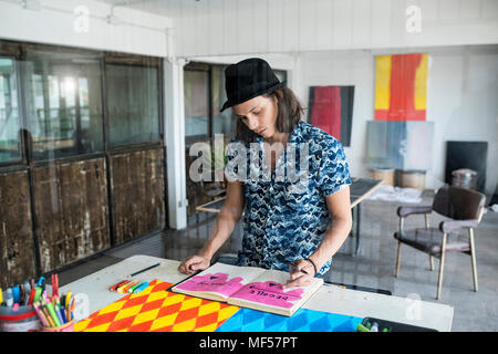 Artista al lavoro, disegno in un notebook nel suo loft studio con dipinti e opere d'arte in background Foto Stock
