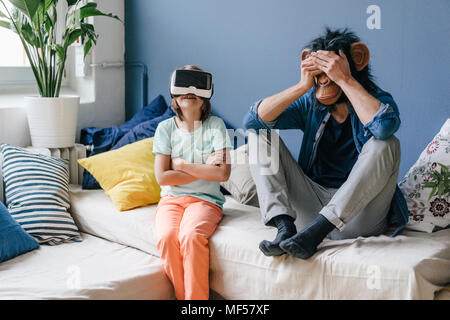 Padre indossa la maschera di scimmia seduto accanto al Figlio che indossa gli occhiali VR a casa Foto Stock