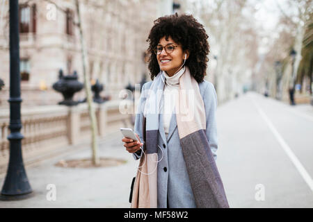 Spagna, Barcellona, Donna sorridente con un telefono cellulare e gli auricolari in città Foto Stock