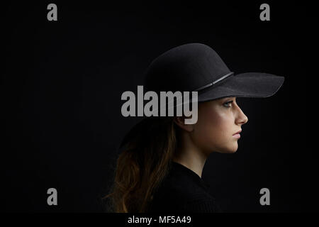 Profilo di triste giovane donna indossa cappello nero su sfondo nero Foto Stock