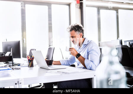 Imprenditore maturo seduto alla scrivania in ufficio utilizzando laptop e di bere il caffè Foto Stock