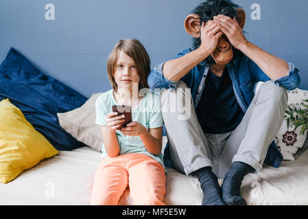 Padre indossa la maschera di scimmia seduto accanto al figlio utilizza lo smartphone a casa Foto Stock