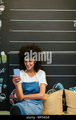 Donna sorridente seduta sul banco in un negozio utilizzando il telefono cellulare Foto Stock