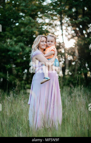 Una donna incinta mentre tiene il suo giovane figlio nelle sue braccia. Un ritratto di un sorridente aspettano mamma e bimbo boy cuddling in un campo al tramonto. Foto Stock