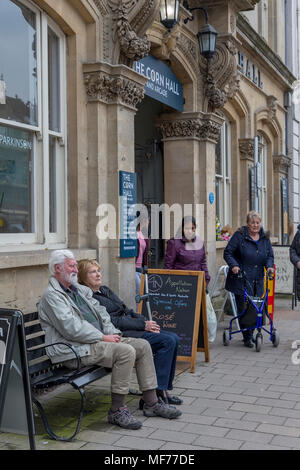 Un paio di rilassarsi seduti su una panchina al di fuori della sala di mais in Cirencester con gli acquirenti di uscire dal porticato Foto Stock