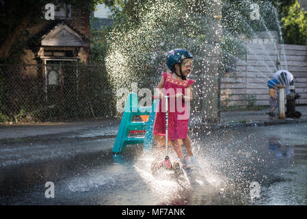 Un bambino di sei anni ragazza con faccia le catture di vernice il sole, equitazione il suo scooter attraverso il getto di un idrante di fuoco in un caldo giorno d'estate. Brooklyn, NY, 2017. Foto Stock