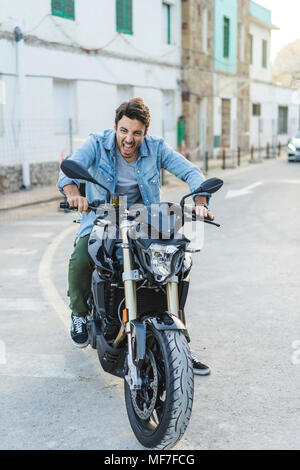 Ritratto di urlando giovane uomo seduto sulla moto tirando funny faces Foto Stock