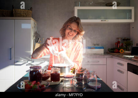 Donna sorridente rendendo confettura di fragole in cucina a casa Foto Stock