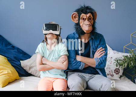 Padre indossa la maschera di scimmia seduto accanto al Figlio che indossa gli occhiali VR a casa Foto Stock