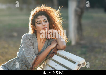 Spagna, Andalusia, Granada. Bella giovane donna con capelli ricci seduta su una panchina in un parco urbano. Il concetto di stile di vita. Foto Stock