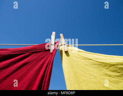 Uno rosso e uno giallo T-shirt appeso su una linea di lavaggio, tenuto con vestiti in legno pioli. Girato a colori dal di sotto, con un cielo blu chiaro dietro. Foto Stock