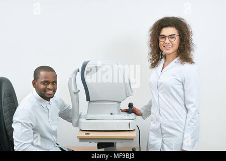 Foto di expirienced, esperti, professinal medico curante sorridendo, soddisfatto di salute del paziente nel gabinetto oculistico. L uomo si trova in prossimità di apparecchiature mediche per la visione del controllo. Foto Stock