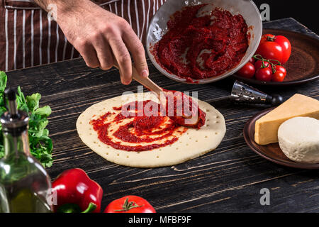 Un maschio di spargimento a mano la salsa di pomodoro sulla base per pizza con cucchiaio su un vecchio sfondo di legno Foto Stock