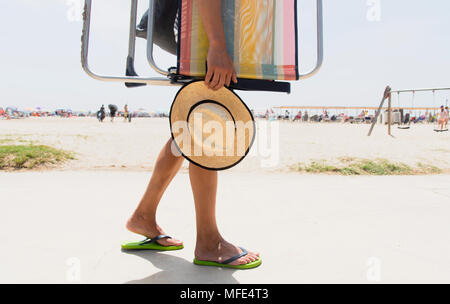 Primo piano di un giovane uomo caucasico portante una sdraio sulla spiaggia e un cappello di paglia in mano, andare e venire dalla spiaggia Foto Stock