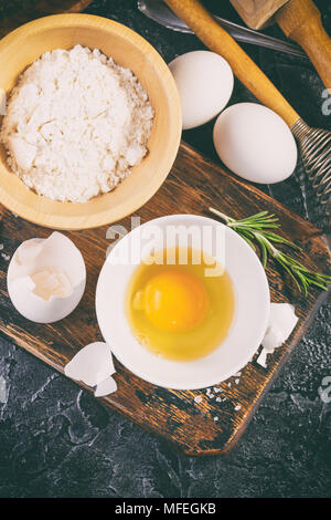 Materie non cotti farina, uova, rosmarino, sale e olio di oliva sul tagliere di legno su sfondo scuro. Foto Stock