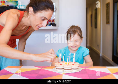Tre anni bambino biondo sorridente guardando la donna accendendo candele con leggero in mano sulla torta di compleanno sulla tovaglia colorata a casa Foto Stock