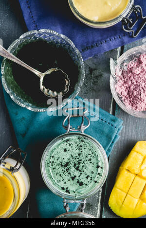 La cheesecake su un vasetto di vetro, fatta con mango e spirulina in polvere, con semi di Chia e polvere di noce di cocco, su una tavola di legno sfondo blu Foto Stock