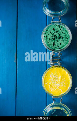 La cheesecake su un vasetto di vetro, fatta con mango e spirulina in polvere, con semi di Chia e polvere di noce di cocco, su una tavola di legno sfondo blu Foto Stock