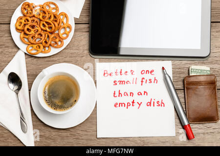 Tovagliolo proverbio del messaggio sul tavolo di legno con caffè, alcuni alimenti e tablet PC meglio di uno piccolo pesce di un piatto vuoto Foto Stock